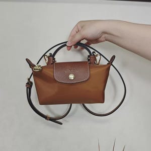 May include: A brown handbag with a leather flap and gold-colored hardware. The bag has a top handle and a detachable shoulder strap. The bag is made of a nylon material.