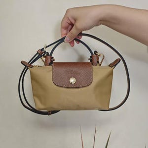 May include: A tan handbag with a brown leather flap and handle. The bag has a gold-colored clasp and a black strap. The bag is held by a hand against a neutral background.