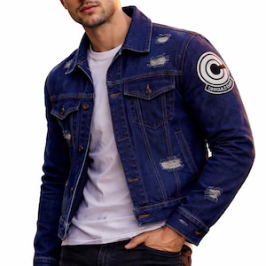 Op de afbeelding: Een spijkerjasje in een medium wassing met distressed details en een Capsule Corp-patch op de bovenarm. De jas heeft een klassieke kraag, manchetten met knopen en borstzakken. De persoon draagt een wit t-shirt en een zwarte jeans.