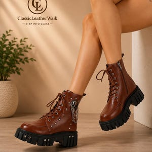 May include: Brown leather ankle boots featuring a lace-up front, side zipper, silver-toned metal studs, and a decorative skull charm with a chain. The boots have a thick, black, lugged rubber sole with silver-toned hardware accents.