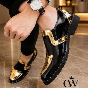 Op de afbeelding: Een paar zwarte en gouden lakleren loafers met een wingtip-ontwerp met decoratieve perforaties, een dikke zwarte profielzool en een glanzende afwerking. De schoenen zijn geaccentueerd met metallic gouden panelen op de neus en zijkant.