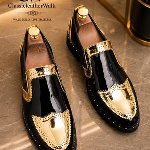 Op de afbeelding: Een paar zwarte lakleren loafers met metallic gouden accenten op de neus en zijpanelen. De schoenen zijn voorzien van geperforeerde details in brogue-stijl langs de gouden randen en hebben houten schoenspanners aan de binnenzijde.
