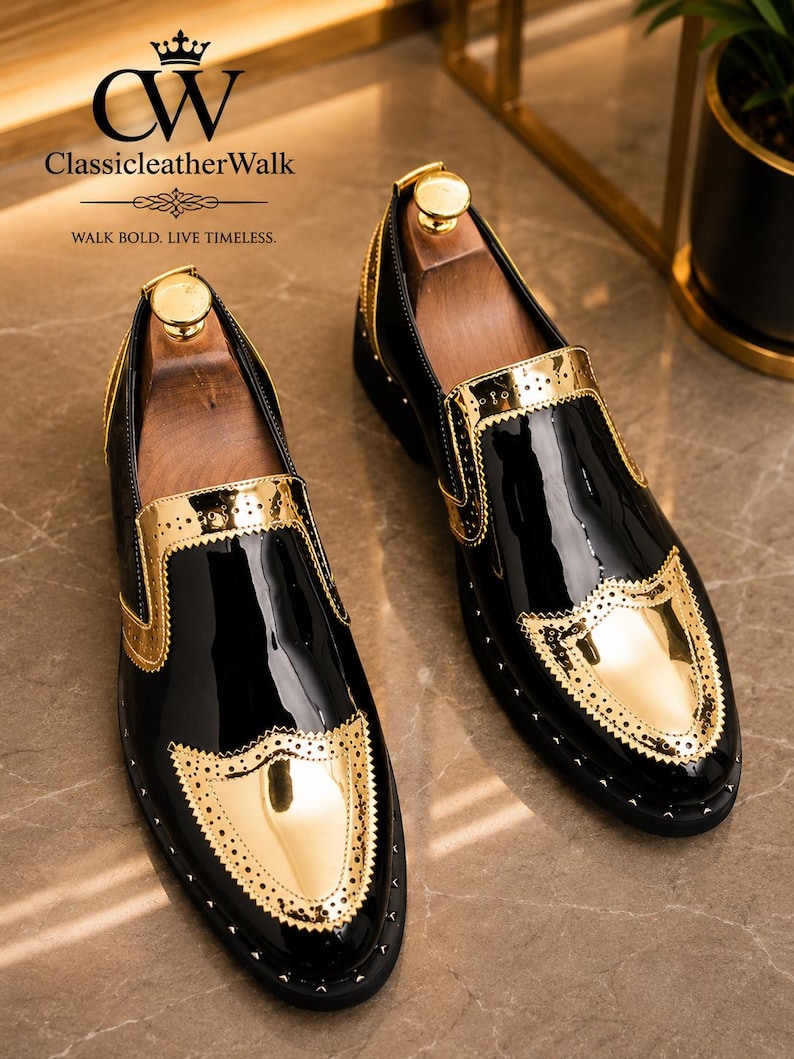 Op de afbeelding: Een paar zwarte lakleren loafers met metallic gouden accenten op de neus en zijpanelen. De schoenen zijn voorzien van geperforeerde details in brogue-stijl langs de gouden randen en hebben houten schoenspanners aan de binnenzijde.