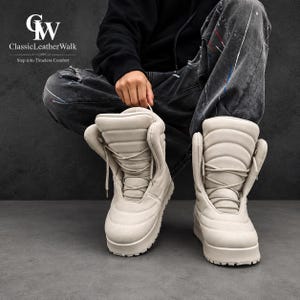 Può includere: Un paio di stivali alti color beige chiaro con un design trapuntato e imbottito, lacci abbinati e una suola spessa e robusta. Il logo "GW ClassicLeatherWalk Step into Timeless Comfort" è visibile nell'angolo in alto a sinistra.