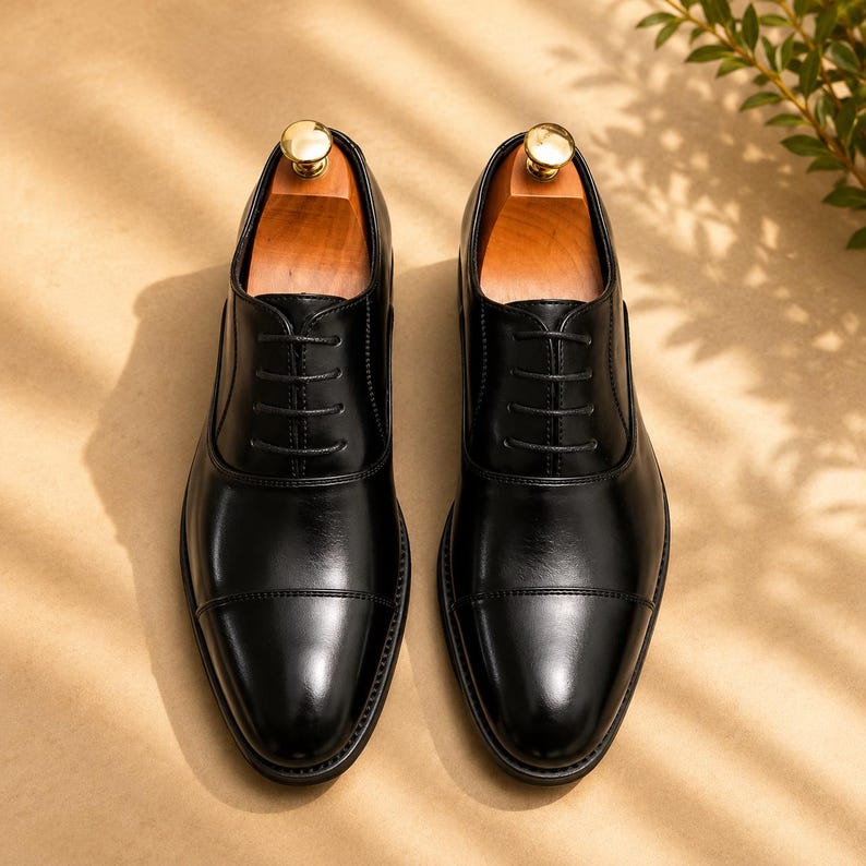 May include: A pair of black leather oxford shoes with a cap toe design, featuring thin black laces and a polished finish. Each shoe includes a light-colored wooden shoe tree inserted inside, with a visible gold-toned metal knob at the heel.