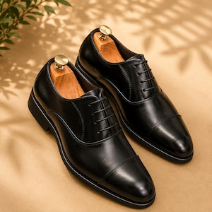 May include: A pair of black leather oxford shoes with a cap toe design, thin black laces, and a smooth finish. Each shoe contains a light-colored wooden shoe tree with a gold-toned metal knob at the heel, showcasing a classic silhouette.