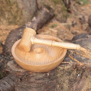 May include: A wooden mortar and pestle set. The mortar is a round bowl with a small indentation in the centre. The pestle is a small mallet. The set is made of light-coloured wood and sits on a tree stump.