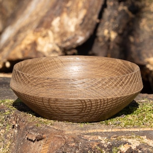 May include: A handcrafted wooden bowl with a natural, light brown finish. The bowl has a wide, shallow design with a textured exterior and a smooth interior. The bowl is sitting on a mossy surface, with a blurred background of wood.