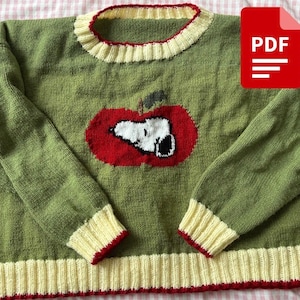 May include: Green knit sweater featuring a Snoopy design inside a red apple. The sweater has cream-colored ribbed cuffs and collar trim with red accents. A PDF icon is in the upper right corner.