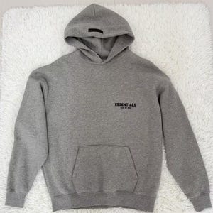 May include: A gray hoodie with a hood and a front pocket. The word "Essentials" is printed in black on the chest. The hoodie is made of a soft material and is suitable for casual wear.
