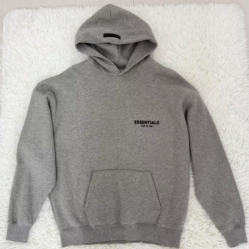 May include: A gray hoodie with a hood and a front pocket. The word "Essentials" is printed in black on the chest. The hoodie is made of a soft material and is suitable for casual wear.