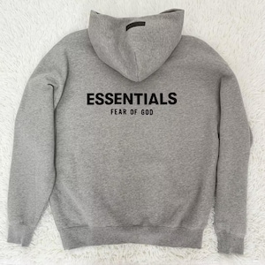 May include: A gray hoodie with the word "ESSENTIALS" printed in black across the chest, with "FEAR OF GOD" printed below. The hoodie has a hood and a small black tag on the back of the hood.