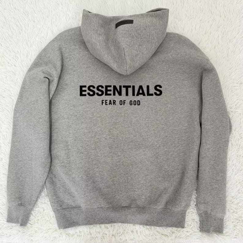 May include: A gray hoodie with the word "ESSENTIALS" printed in black across the chest, with "FEAR OF GOD" printed below. The hoodie has a hood and a small black tag on the back of the hood.
