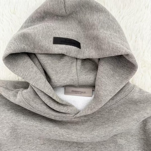 May include: A gray hoodie with a hood and a kangaroo pocket. The hoodie has a black rectangular label on the hood and a rectangular label on the front with the word "ESSENTIALS". The hoodie is made of a soft, comfortable material.