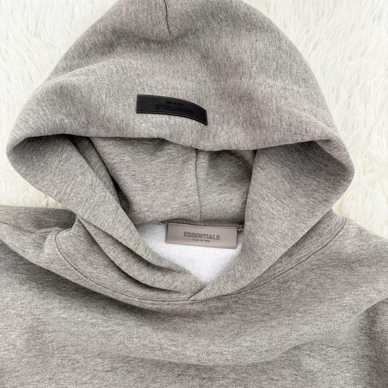 May include: A gray hoodie with a hood and a kangaroo pocket. The hoodie has a black rectangular label on the hood and a rectangular label on the front with the word "ESSENTIALS". The hoodie is made of a soft, comfortable material.