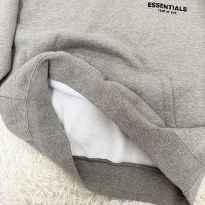 May include: A gray hoodie with the word "ESSENTIALS" printed in black on the upper chest. The hoodie has a white interior lining and a kangaroo pocket. The fabric appears soft and comfortable.