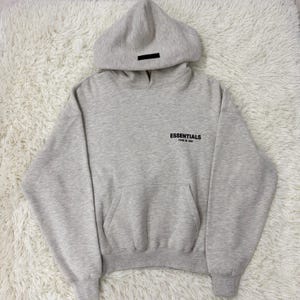 May include: A light gray hoodie with a hood and a front pocket. The word "ESSENTIALS" is printed on the chest in black. A black rectangular patch is on the hood.