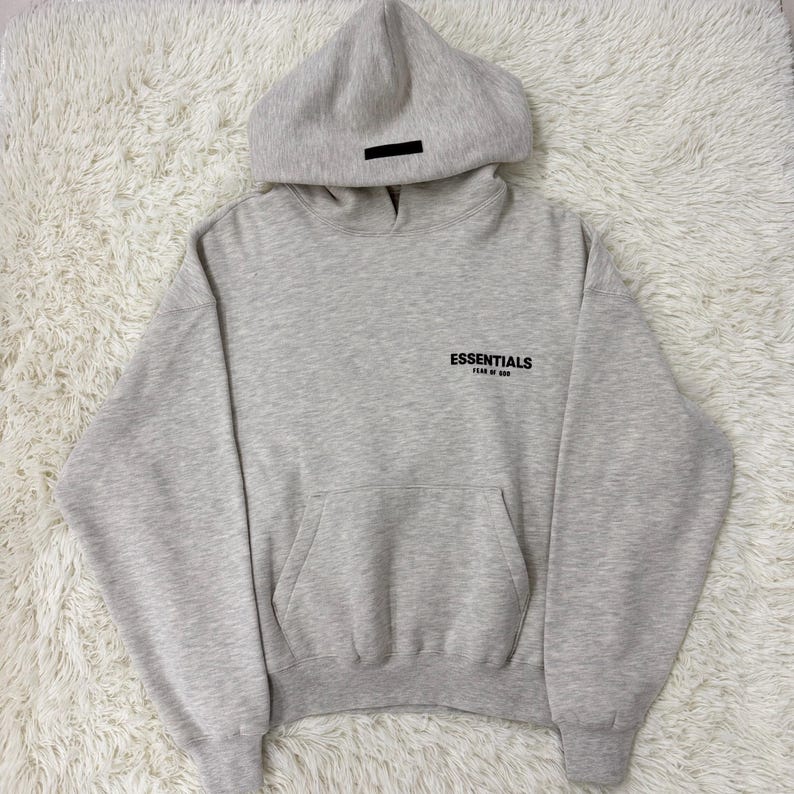 May include: A light gray hoodie with a hood and a front pocket. The word "ESSENTIALS" is printed on the chest in black. A black rectangular patch is on the hood.
