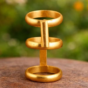 May include: A gold-colored ring holder with three circular bands connected by a vertical bar. The ring holder is displayed on a brown surface with a blurred green and yellow background.