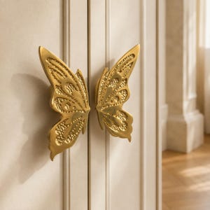 May include: Two gold-colored metal cabinet pulls shaped like butterfly wings, mounted on a light-colored paneled door. Each pull features a polished finish with textured, recessed details within the wing patterns, creating a symmetrical design.