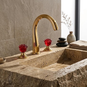 May include: A gold-coloured bathroom tap with a curved spout and two red rose-shaped handles. The tap is installed on a stone sink. The sink has a natural, textured appearance. The overall design is elegant and decorative.