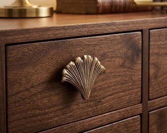 Könnte beinhalten: Nahaufnahme einer Holzschublade mit einem messingfarbenen Griff in Form eines Ginkgoblattes. Die Schublade ist aus dunkelbraunem Holz gefertigt. Der Griff ist ein dekoratives Element. Das Möbelstück ist Teil eines Möbelsets.