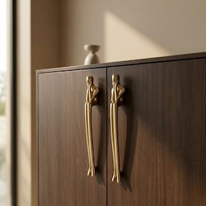 May include: A dark brown wooden cabinet with two doors, each featuring a gold-colored handle in the shape of a stylized human figure. The handles are vertically oriented, adding a unique design element to the furniture.