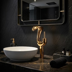 Puede incluir: Una escena de baño moderno con un grifo dorado con un diseño curvo único. Un lavabo redondo blanco se encuentra debajo del grifo. El baño tiene una encimera de mármol negro, un espejo negro y una pared negra. También se ven una toalla negra y un dispensador de jabón.
