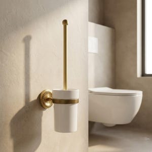 Puede incluir: Un portacepillos de inodoro montado en la pared con una taza de cerámica blanca y un mango y detalles de metal dorado. El soporte se fija a la pared con una base circular decorativa. El cepillo está dentro de la taza.