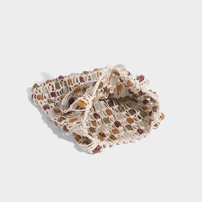 May include: A beige crocheted bag with a grid-like pattern, filled with various-colored stones. The stones are in shades of brown, orange, and white. The bag is folded over, revealing the interior and the stone contents.