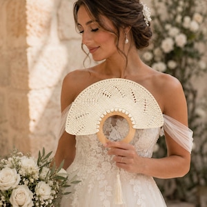 Può includere: Un ventaglio color crema, tessuto, con manico in legno e nappina. Il ventaglio è tenuto davanti a una sposa che indossa un abito da sposa in pizzo bianco. La sposa tiene anche un bouquet di rose bianche.