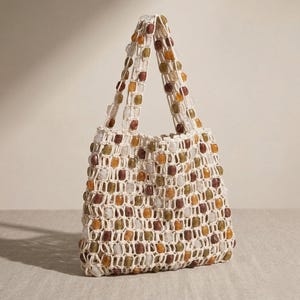 May include: A handmade tote bag with a cream-colored crocheted net design. The bag is adorned with square-shaped beads in shades of brown, orange, green, and white. The handles are also beaded. This stylish bag is perfect for carrying groceries or other items.