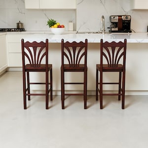 Pode incluir: Três banquetas de bar em madeira castanho-escura com um rebordo superior recortado e um encosto em forma de leque. Cada banco possui quatro pernas direitas, um assento quadrado e barras de suporte horizontais em todos os lados da estrutura.