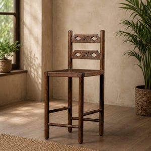 May include: A dark brown wooden bar stool with a square seat and a backrest featuring diamond-shaped cutouts. The stool has a sturdy, rustic design, with visible wood grain and a footrest. The background includes a window, plants, and a neutral-toned wall.