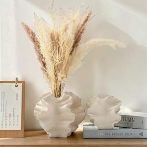 May include: A white, textured ceramic vase with a ruffled design, holding dried pampas grass and a small white flower. A smaller matching vase sits beside it. The arrangement is on a wooden surface.