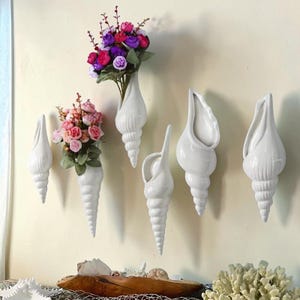 Może przedstawiać: Białe ceramiczne wazony ścienne w kształcie muszli, niektóre wypełnione kolorowymi kwiatami. Kwiaty są w odcieniach różu, fioletu i czerwieni. Wazony są zamontowane na ścianie, tworząc dekoracyjną ekspozycję. U dołu drewniany element dekoracyjny.