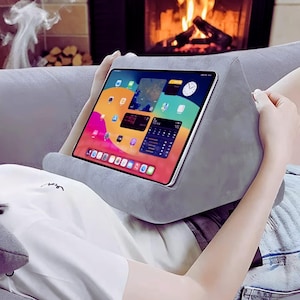 Puede incluir: Una almohada gris para tablet con una tablet que muestra una interfaz colorida. La almohada está diseñada para sostener una tablet mientras se usa. La persona lleva una camiseta blanca y pantalones cortos de mezclilla.