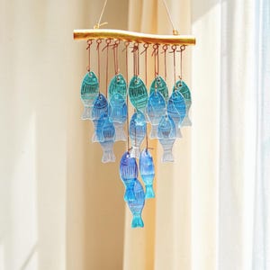 Puede incluir: Un carillón decorativo con adornos en forma de pez en tonos azules y turquesas. Los peces están suspendidos de una barra de madera con alambre color cobre. El carillón está diseñado para colgar.