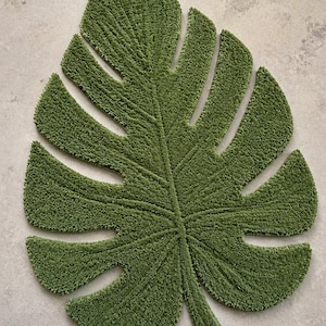 Może przedstawiać: Zielona mata łazienkowa w kształcie liścia. Mata została zaprojektowana tak, aby przypominać liść rośliny Monstera, z teksturowaną, pluszową powierzchnią. Kolor maty to żywa, naturalna zieleń, umieszczona na neutralnej powierzchni.