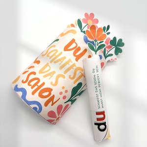 Könnte beinhalten: Eine zylindrische Papierrolle mit blumenförmigem Ausschnitt am oberen Rand, bedruckt mit dem Text "DU SCHAFFST DAS SCHON" in Orange und Gelb. Eine kleinere weiße Papierrolle ist befestigt, bedruckt mit "DU SCHAFFST DAS SCHON. DU MUSST NICHT PERFEKT SEIN." und "du".