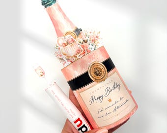 Può includere: Una sagoma decorativa a forma di bottiglia di champagne con un design ad acquerello rosa e crema, una fascia nera, un sigillo dorato e il testo Happy Birthday, Ich wünsche dir nur das Allerbeste. Un piccolo tubetto con il testo BIST HEUTE DER GRUND ZUM FEIERN è attaccato.