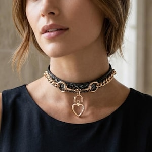 May include: A black textured choker necklace featuring a gold-toned chain link design and a central gold-toned heart-shaped pendant. The choker is constructed with a dark, snakeskin-patterned band and metallic hardware connecting the chain to the pendant.