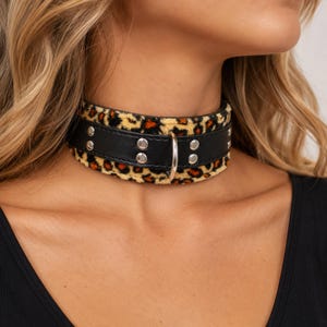 May include: A black and leopard print choker necklace. The choker features a black leather band with silver rivets and a D-ring. The leopard print fabric is tan, brown, and black.