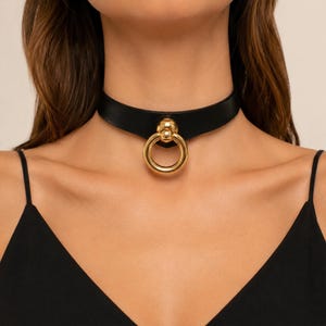 May include: Black choker necklace with a large gold ring pendant. The choker is made of a smooth, black material and fastens around the neck. The gold ring is a prominent feature, adding a bold accent to the design. The necklace is a fashion accessory.