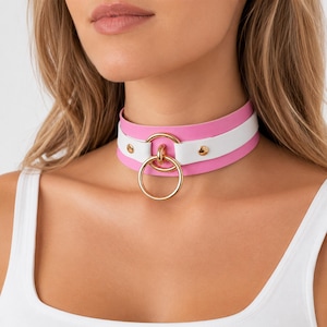 May include: A pink, white, and gold choker necklace. The choker features a pink band, a white band, and a gold ring. The choker is secured with gold-colored rivets.