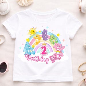 Peut inclure: T-shirt blanc avec un motif coloré de Bisounours, un arc-en-ciel et le chiffre 2. Le texte "Birthday Girl" est écrit en dessous. Le t-shirt est entouré d'accessoires, dont des chaussures, des lunettes de soleil et un sac à main.