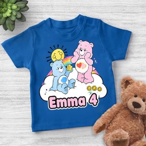 Peut inclure: T-shirt bleu pour enfant avec un motif de deux Bisounours sur un nuage, un arc-en-ciel et un soleil. Le nom "Emma 4" est imprimé en rose. Un ours en peluche marron est visible en arrière-plan.
