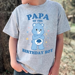 Peut inclure: T-shirt gris avec un ours bleu de dessin animé et le texte "PAPA of the BIRTHDAY BOY". L'ours a un motif de nuage sur le ventre et est entouré d'étoiles et de cœurs colorés.