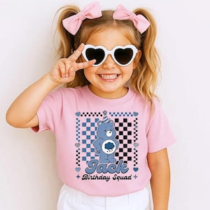 Peut inclure: Un t-shirt rose avec un motif d'ours bleu portant un chapeau de fête, sur un motif à carreaux. Le texte "Jack Birthday Squad" est en dessous de l'ours. La fille porte des lunettes de soleil en forme de cœur et des nœuds roses.