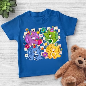 Peut inclure: Un t-shirt bleu avec un motif de quatre oursons de dessin animé portant des chapeaux de fête, sur un fond à carreaux avec des fleurs et des étoiles. Les ours sont violets, verts, bleus et jaunes. Un ours en peluche marron est à droite.